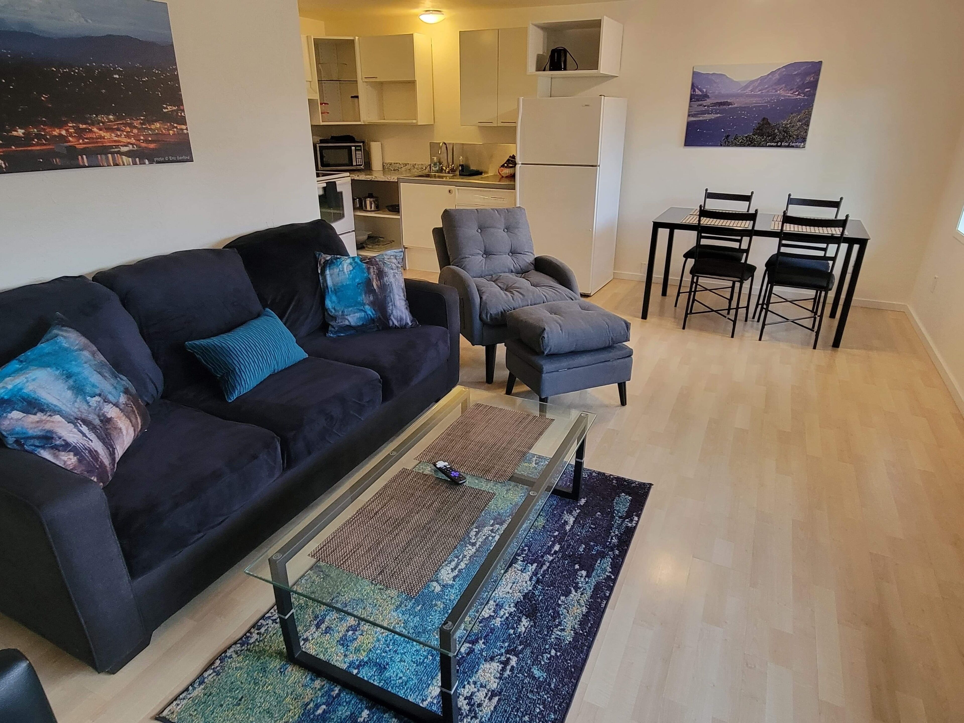 A living room with a dark sofa, a glass coffee table, and a patterned rug. There is a kitchen in the background with white cabinets and a refrigerator.