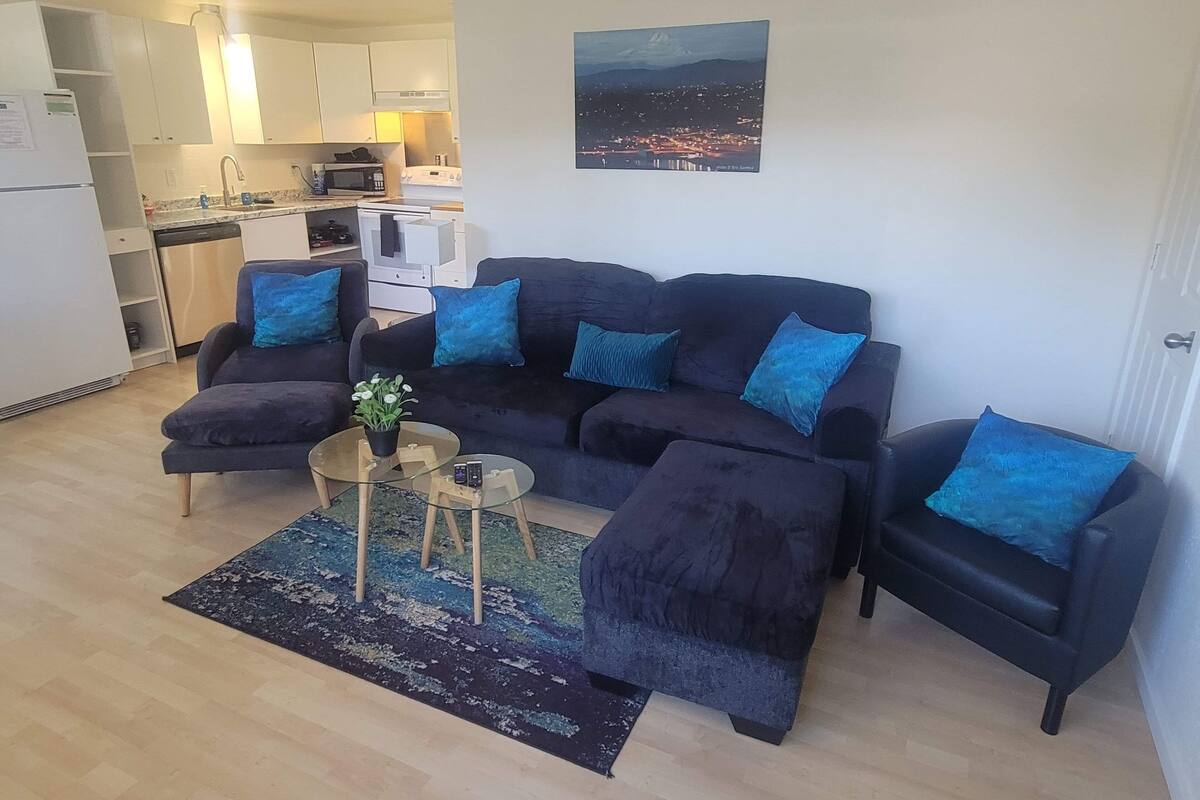 Una sala de estar con un sofá azul oscuro, una mesa de centro de vidrio y una mesita auxiliar con una planta. Se ve una cocina al fondo con armarios blancos y un microondas.