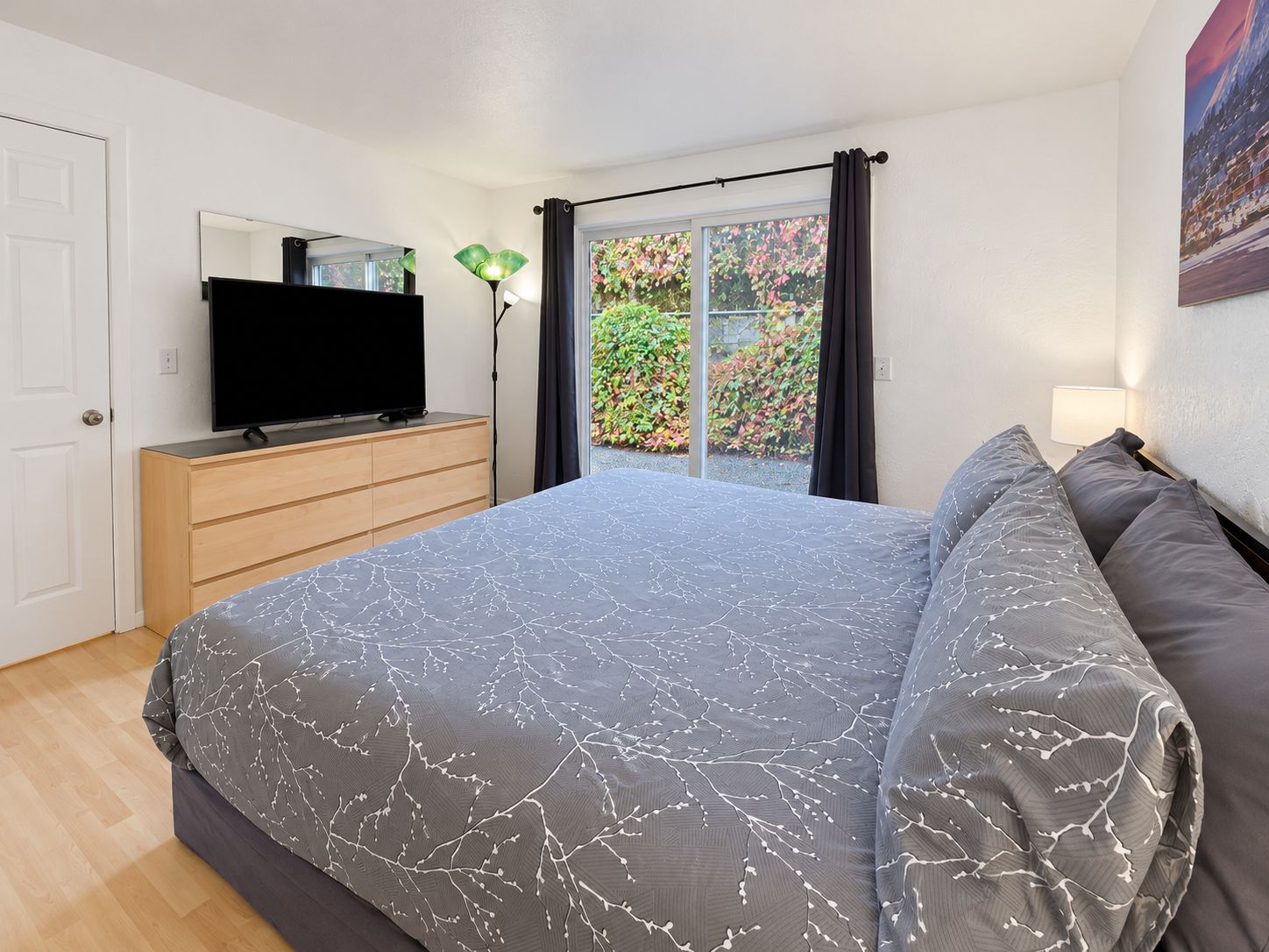 A bedroom with a bed, a TV on a dresser, a window with curtains, and a painting on the wall.
