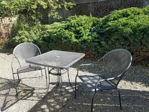 Une table en métal et deux chaises, disposées dans un espace extérieur, avec un sol de gravier et un fond de verdure.