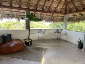 A room with a thatched roof, a large window, a sofa, two bar stools, and a potted plant.