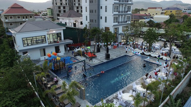 A swimming pool area with a water slide and lounge chairs, surrounded by buildings and greenery. - Romano Hotel (Kampot)