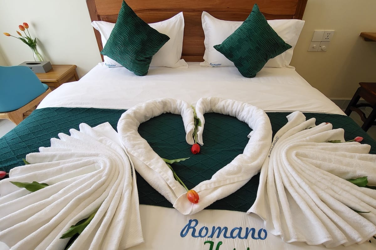 A neatly made bed with a heart-shaped towel arrangement and a rose, and the text "Romano Hotel" on the bedspread.