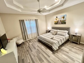 A bedroom with a large bed, a ceiling fan, a window with curtains, a bedside table with a lamp, and a painting on the wall.
