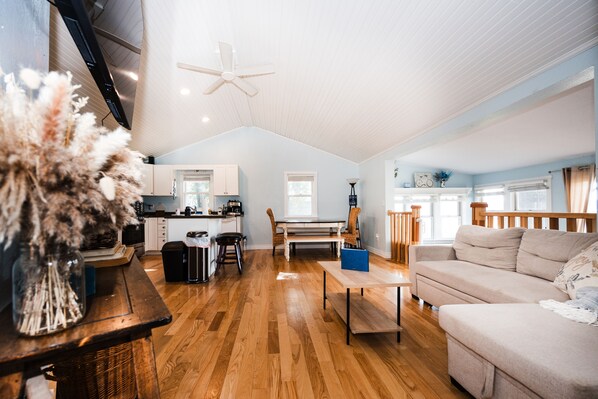 A spacious living room with a wooden floor, a sofa, a dining table, and a kitchen area in the background.