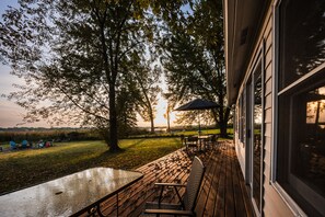 An outdoor deck with a table and chairs, overlooking a grassy area with trees and a sunset in the background.
