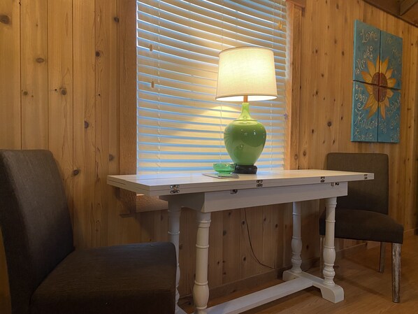 A wooden interior with a white table, two chairs, a green lamp, and a sunflower painting.