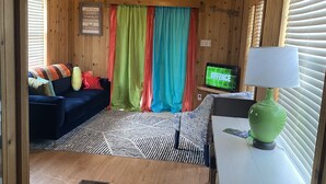 L’intérieur d’un chalet accueillant, avec un plafond en bois, un canapé bleu, des rideaux colorés, un téléviseur et une lampe verte.