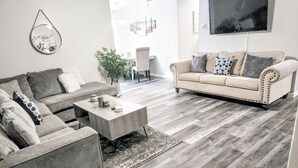 Un salon avec un canapé gris, un canapé beige, une table basse et un téléviseur fixé au mur.