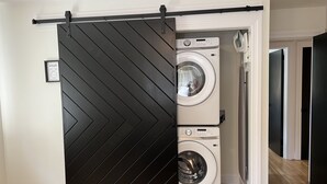 A modern laundry room with a large black chevron-patterned sliding door, two stacked front-loading washing machines, and a framed 'wash' sign on the wall. - Restful Mid-Mod in the heart of Hickory (Hickory)