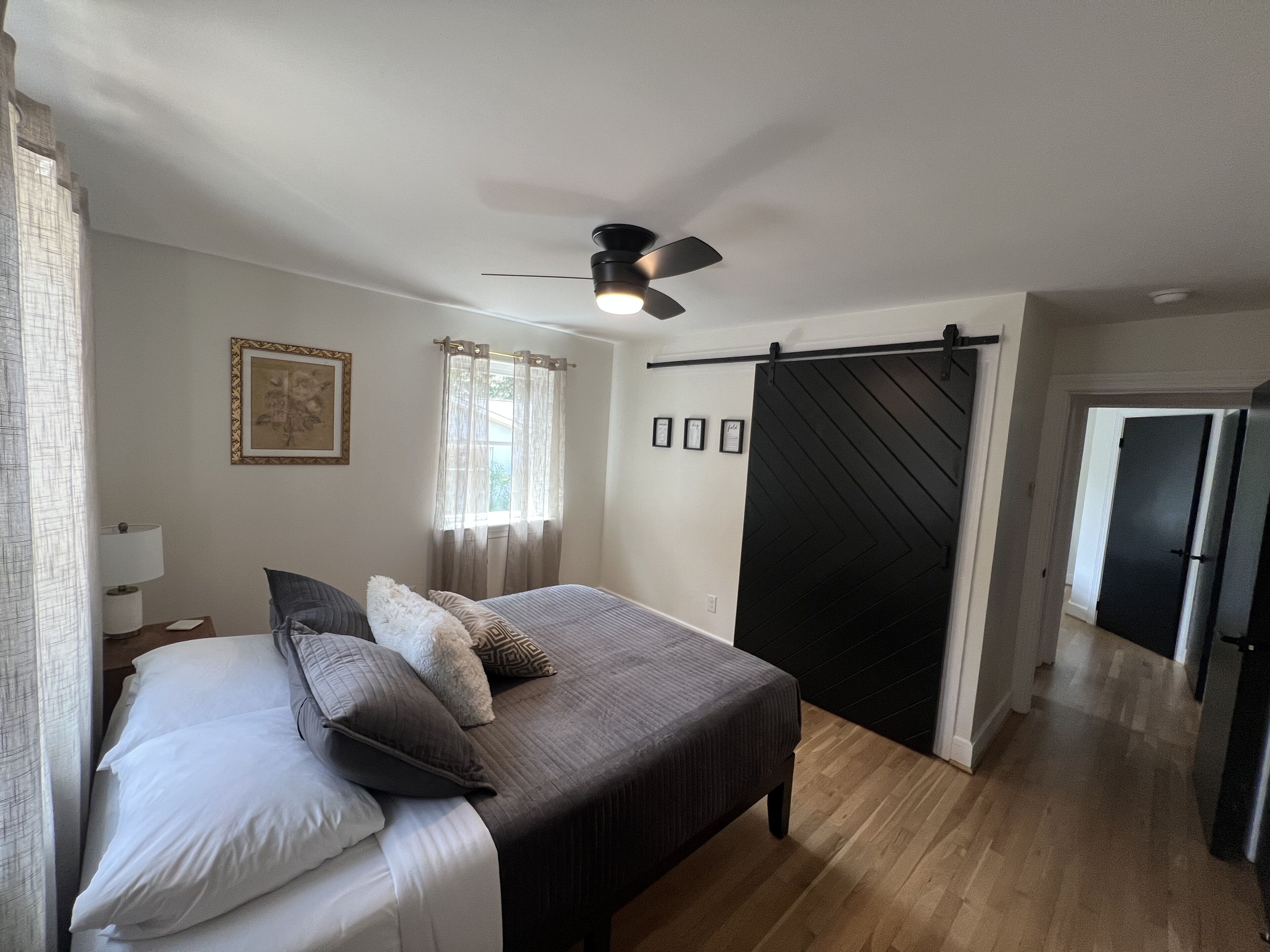 Ein Schlafzimmer mit Bett, Deckenventilator, Fenster mit Vorhängen, einer Tür und einem Flur.