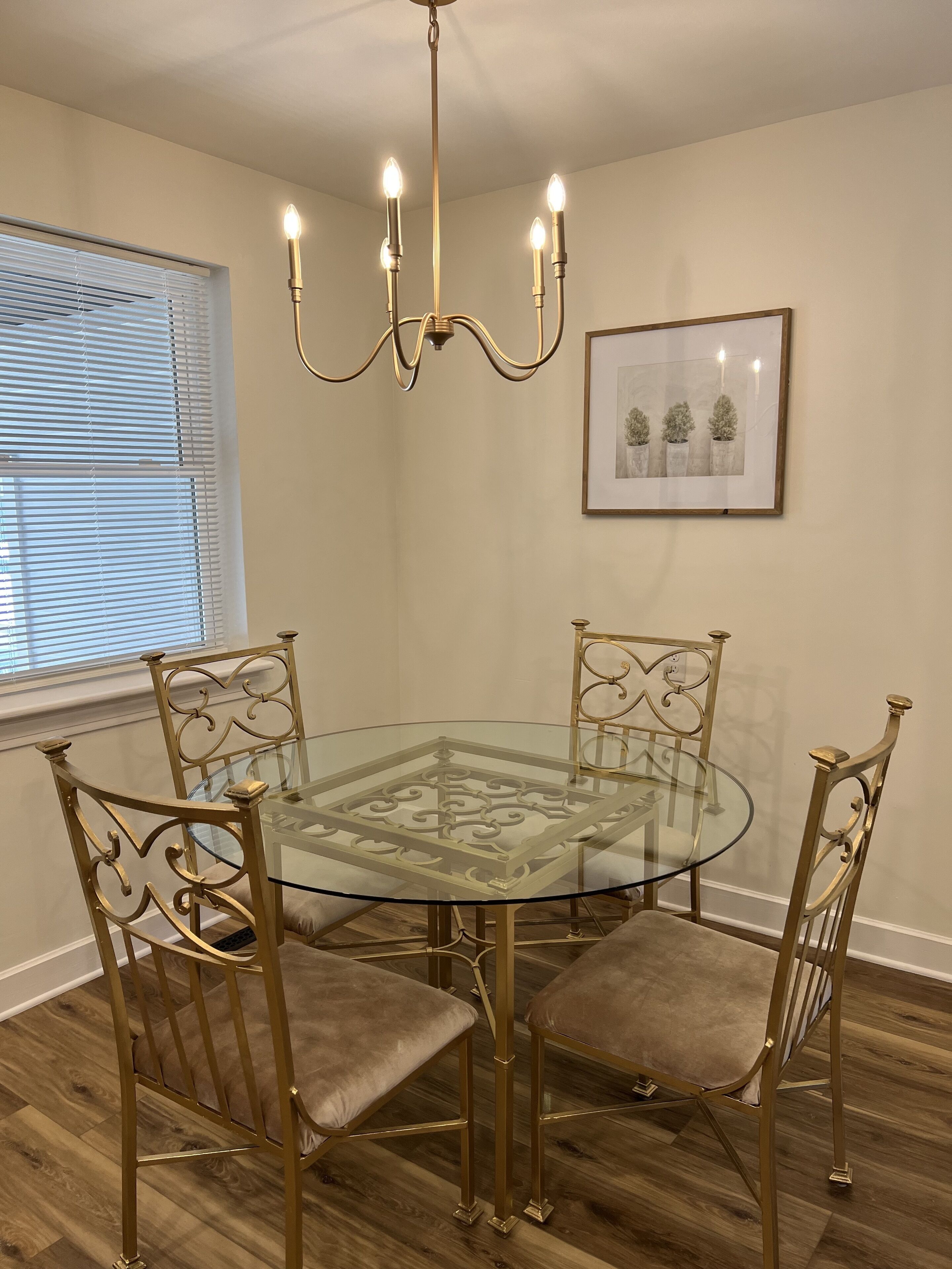 A dining area with a round glass table, four upholstered chairs, a chandelier, and a framed picture on the wall.