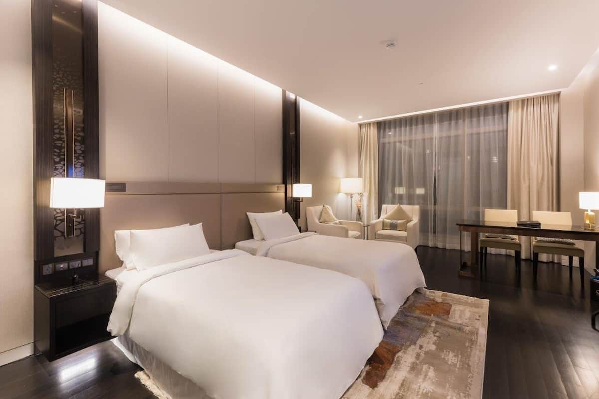 Habitación de hotel con dos camas, un escritorio, sillas y un ventanal con cortinas.