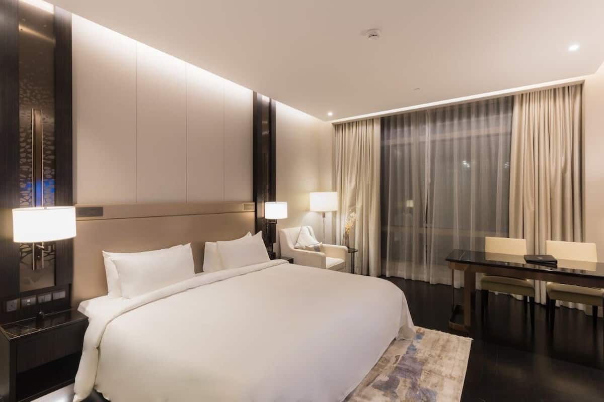 Una habitación de hotel con una cama grande, mesitas de noche con lámparas, un escritorio y un amplio ventanal con cortinas.