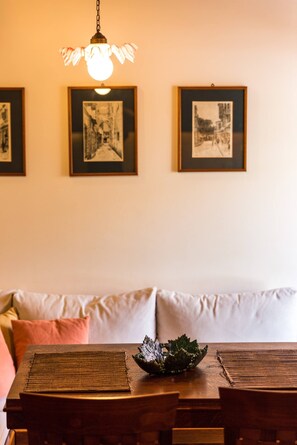 A dining area with a wooden table, a woven placemat, and a decorative bowl. There are framed pictures on the wall and a hanging light fixture.