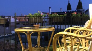 En balkon med fletmøbler, udsigt over bybilledet og et blomsterprydet hegn.