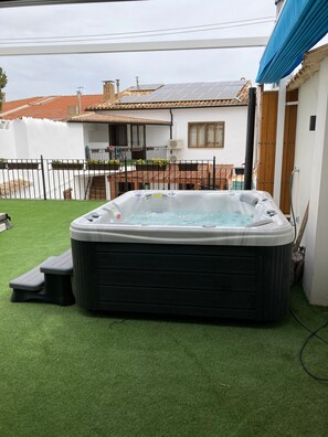 A hot tub in a backyard with artificial grass, a wooden deck, and a white house with a tiled roof.