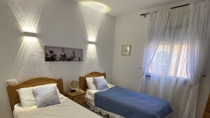Une chambre avec deux lits, une table de chevet, une lampe fixée au mur, une fenêtre avec des rideaux et un tableau accroché au mur.