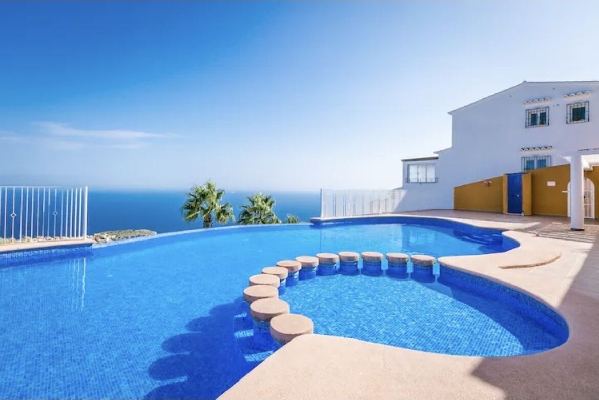 A swimming pool with a curved edge, a clear blue sky, and a building with a white facade and yellow accents in the background.