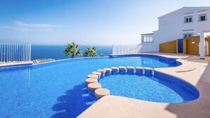 Une piscine à bord incurvé, un ciel bleu azur et, au fond, un bâtiment à la façade blanche rehaussée de touches de jaune.