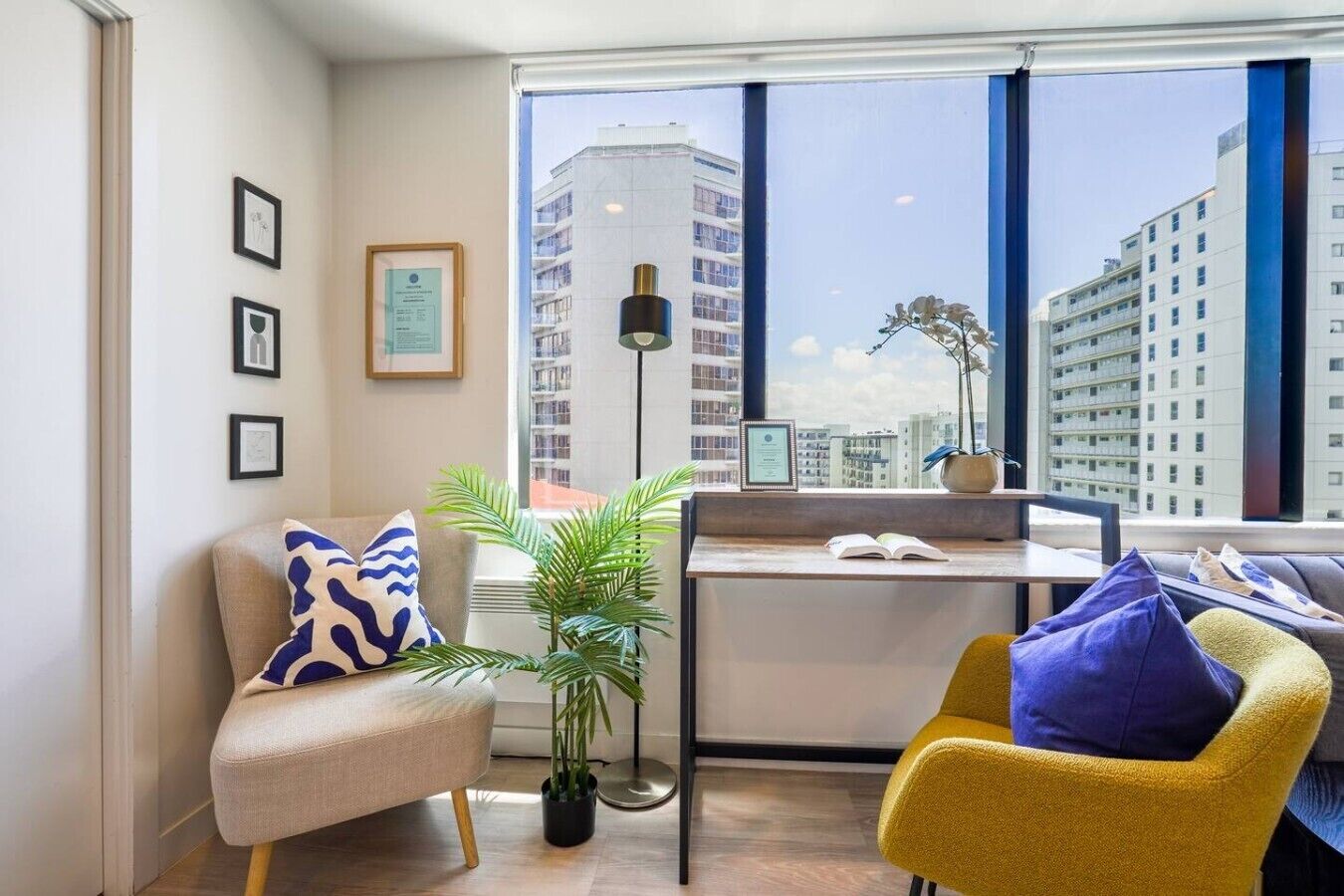 A modern living room with a sofa, a yellow chair, a small table, and a large window with a city view.