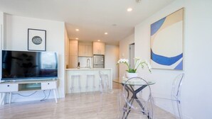 A modern living room with a flat-screen TV, a glass dining table with chairs, a kitchen in the background, and a large abstract painting on the wall. - Two Bedroom Apartment Right Next to the Sky Tower (Auckland)