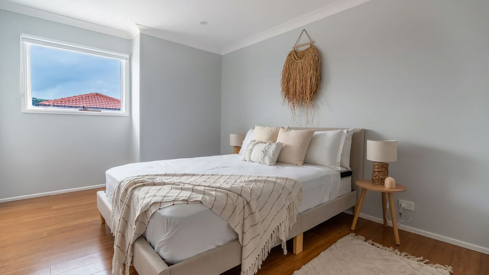 A bedroom with a bed, bedside lamps, a hanging decor, and a window with a view of a red-roofed building.