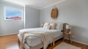 A bedroom with a bed, bedside lamps, a hanging decor, and a window with a view of a red-roofed building.