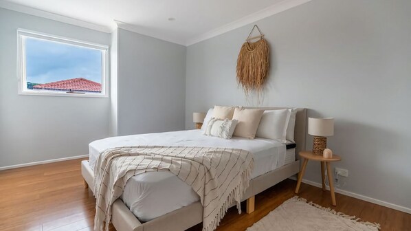 A bedroom with a bed, bedside lamps, a hanging decor, and a window with a view of a red-roofed building.