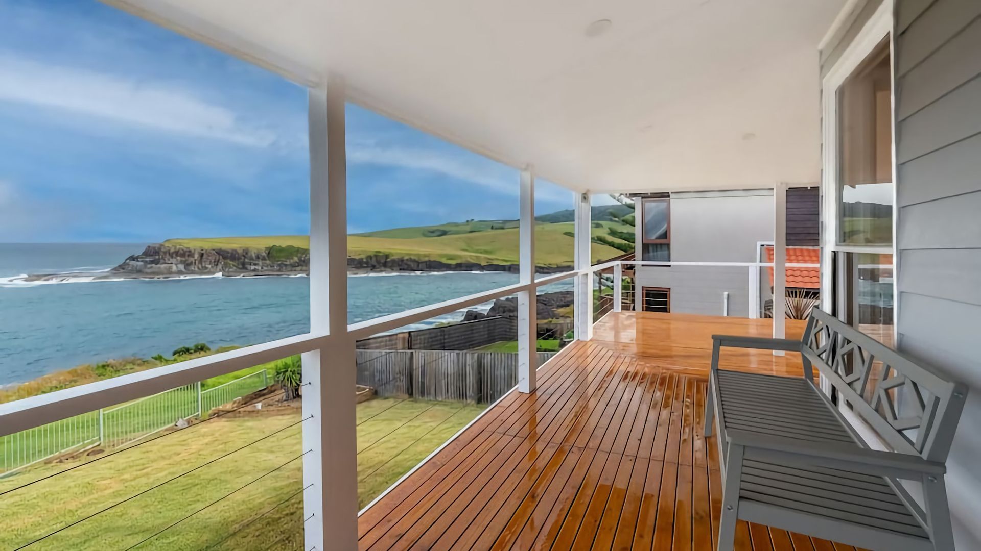 A balcony with a wooden floor, white railings, and a view of the ocean and hills.