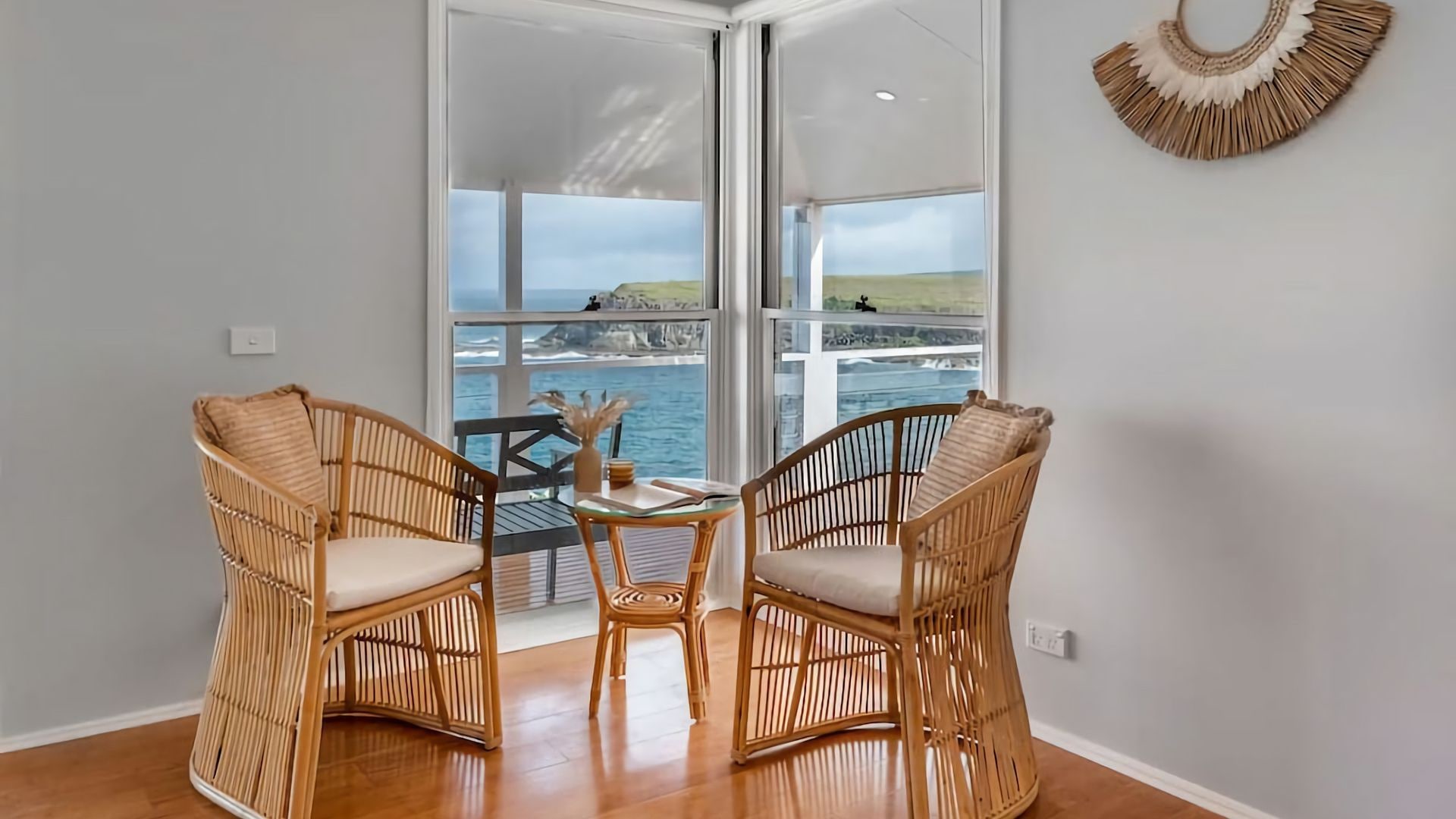 A room with two wicker chairs, a small table, and a view of the sea through a glass door.