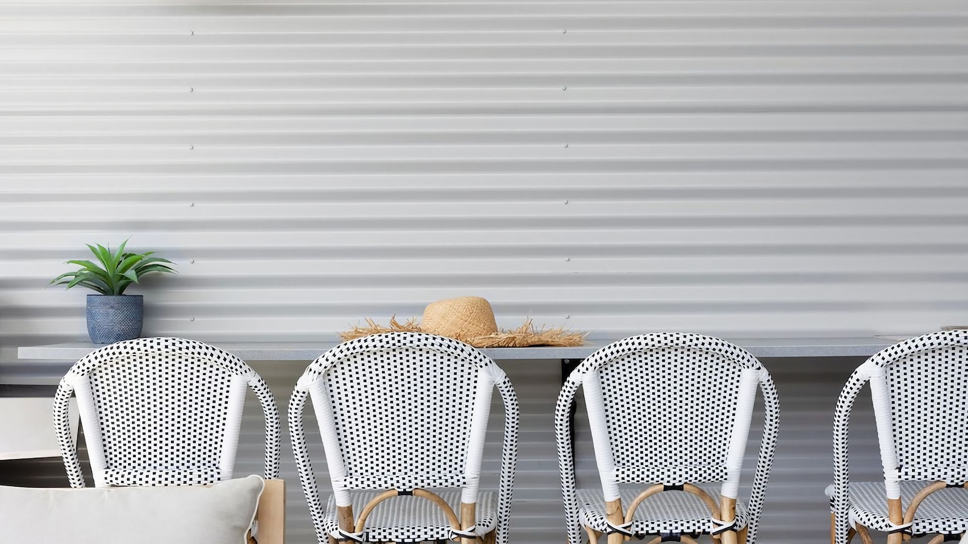 A row of white chairs with black checkered patterns, a potted plant, and a straw hat on a table against a corrugated metal wall.