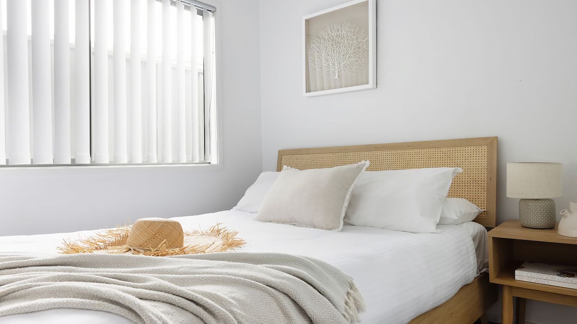 A bedroom with a bed, a bedside table with a lamp, a window with blinds, and a framed artwork on the wall.