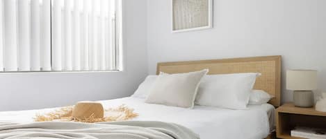 Un dormitorio con cama, una mesita de noche con lámpara, una ventana con persianas y una obra de arte enmarcada en la pared.