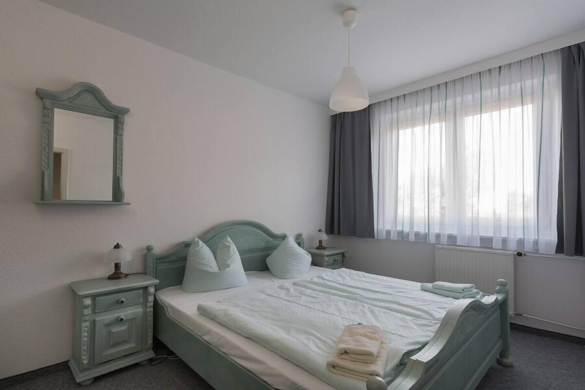 Une chambre à coucher avec un lit, une table de chevet, un miroir et une fenêtre avec des rideaux.