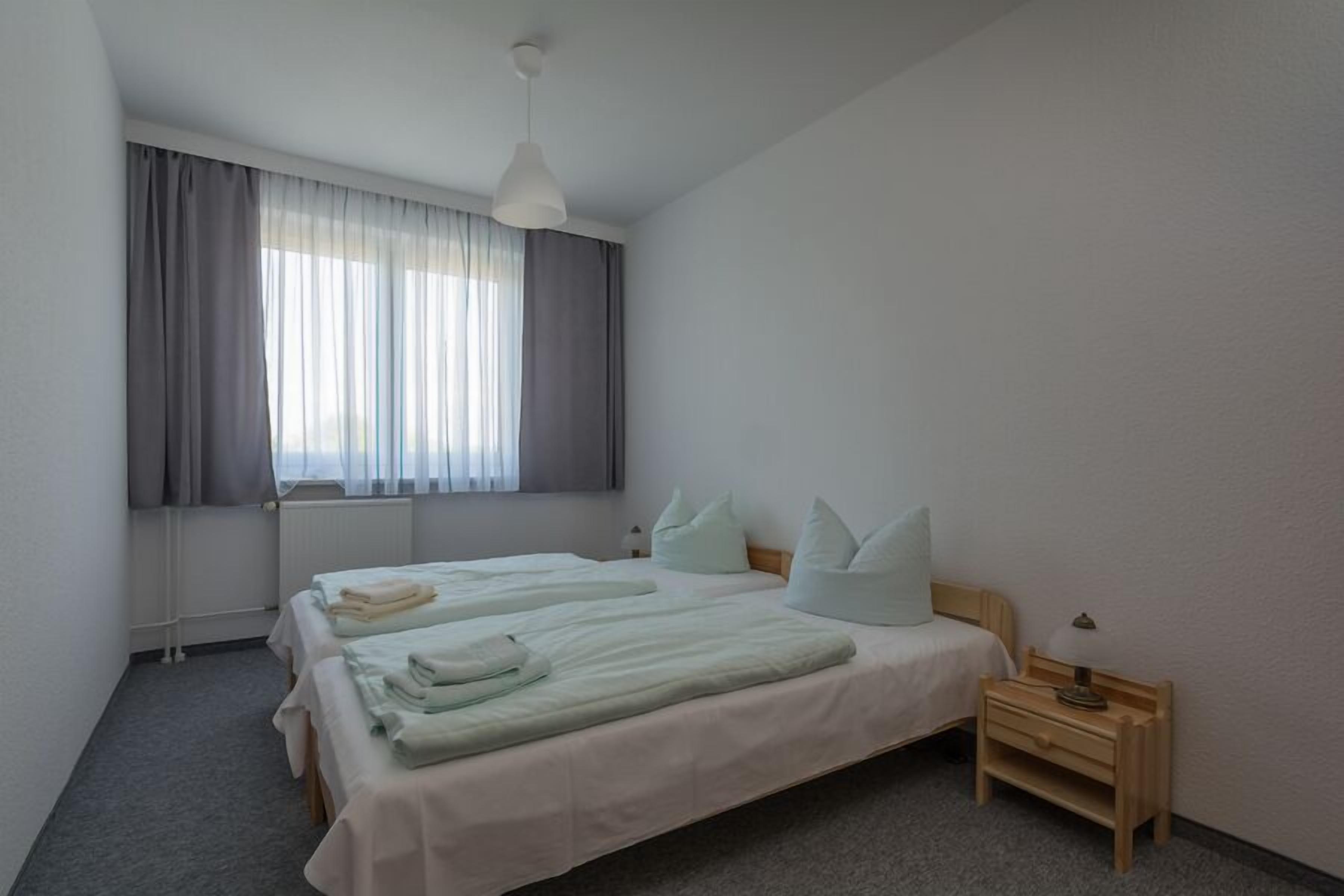Una habitación de hotel con dos camas, cortinas grises, una lámpara de techo blanca y una mesita de noche de madera con lámpara.