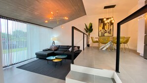 Un salon moderne avec un canapé d’angle noir, une table basse et un coin repas avec des chaises jaunes. Un escalier mène à un autre niveau, et une grande fenêtre est dotée de stores.