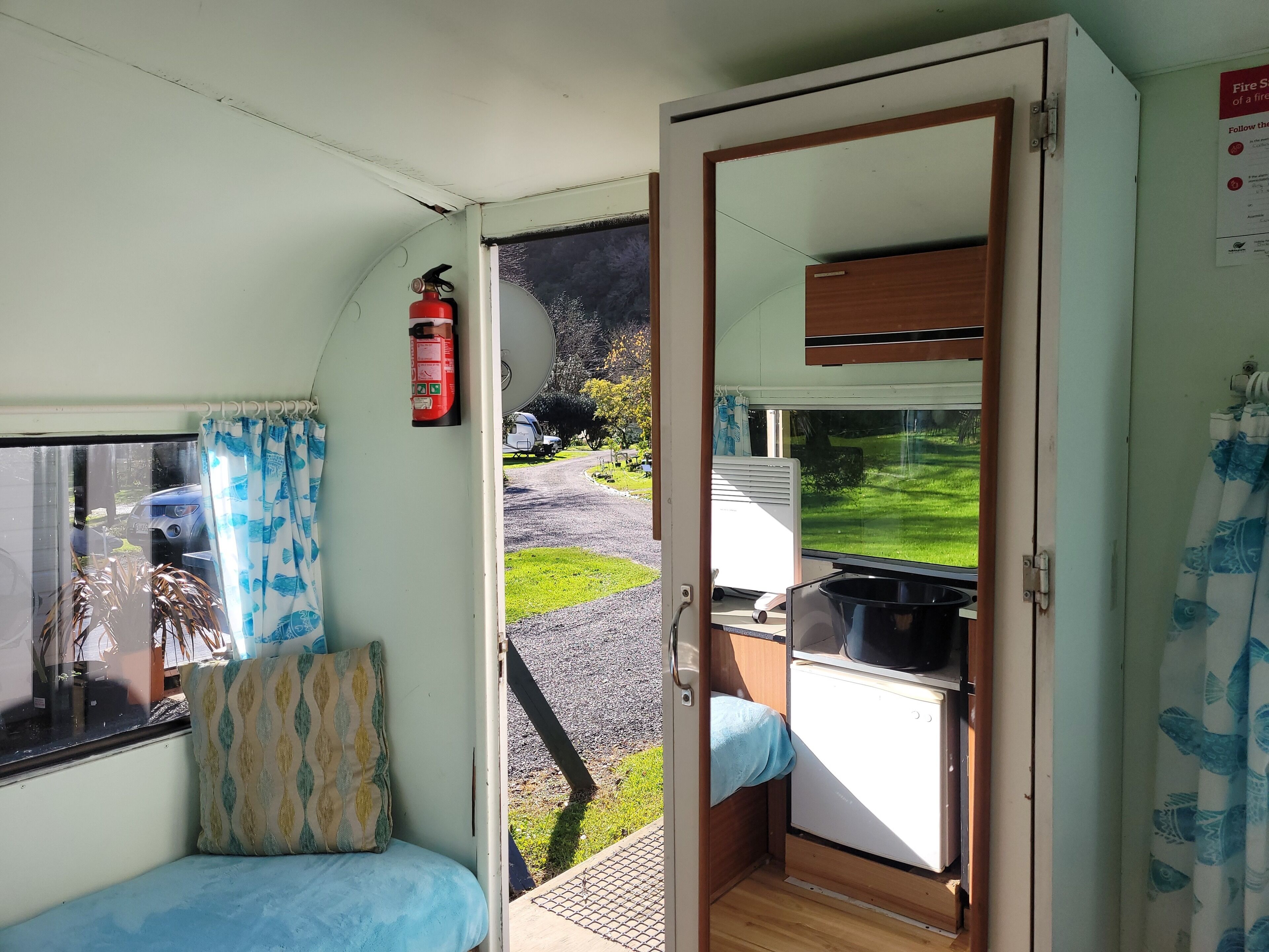Interior compacto de una caravana con kitchenette, zona de descanso con cojines estampados y una puerta que da al exterior.