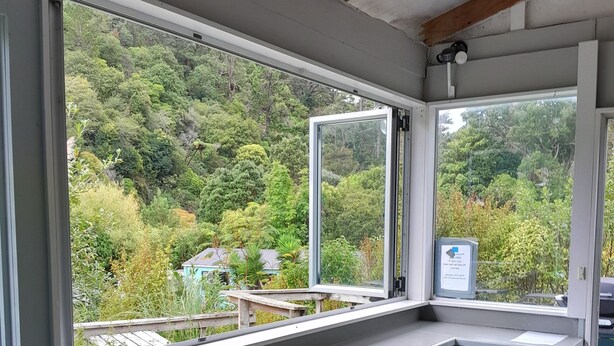 A room with a large window overlooking a forested area, a white desk with black stools, and a small sign on the window.