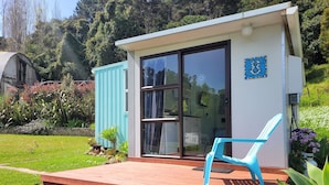 A small, modern house with a glass door, a blue chair on the porch, and a garden with various plants.