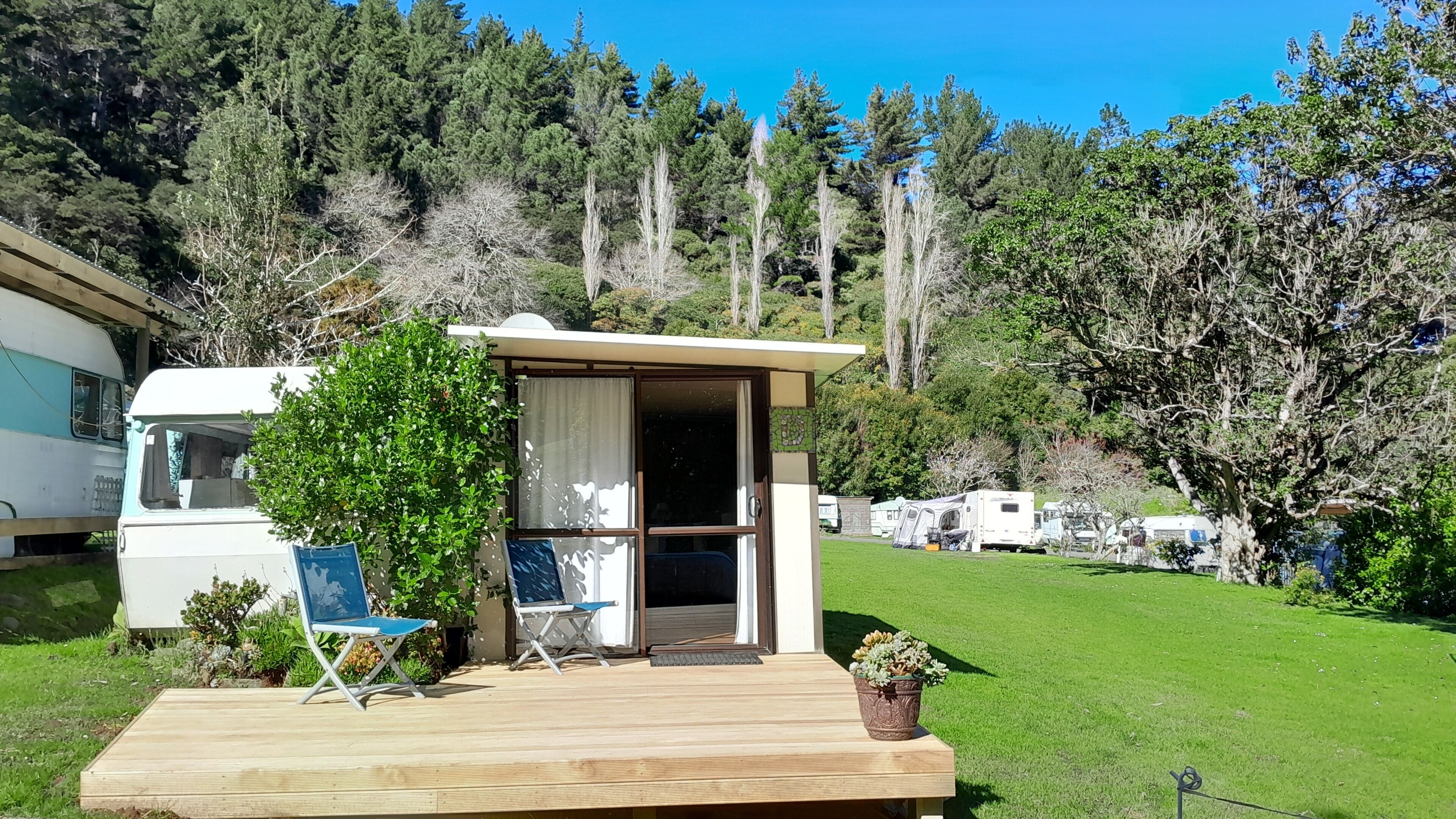 Eine kleine Holzterrasse mit zwei Stühlen, einem Topfpflanzen und einem Gebäude mit Glastür.