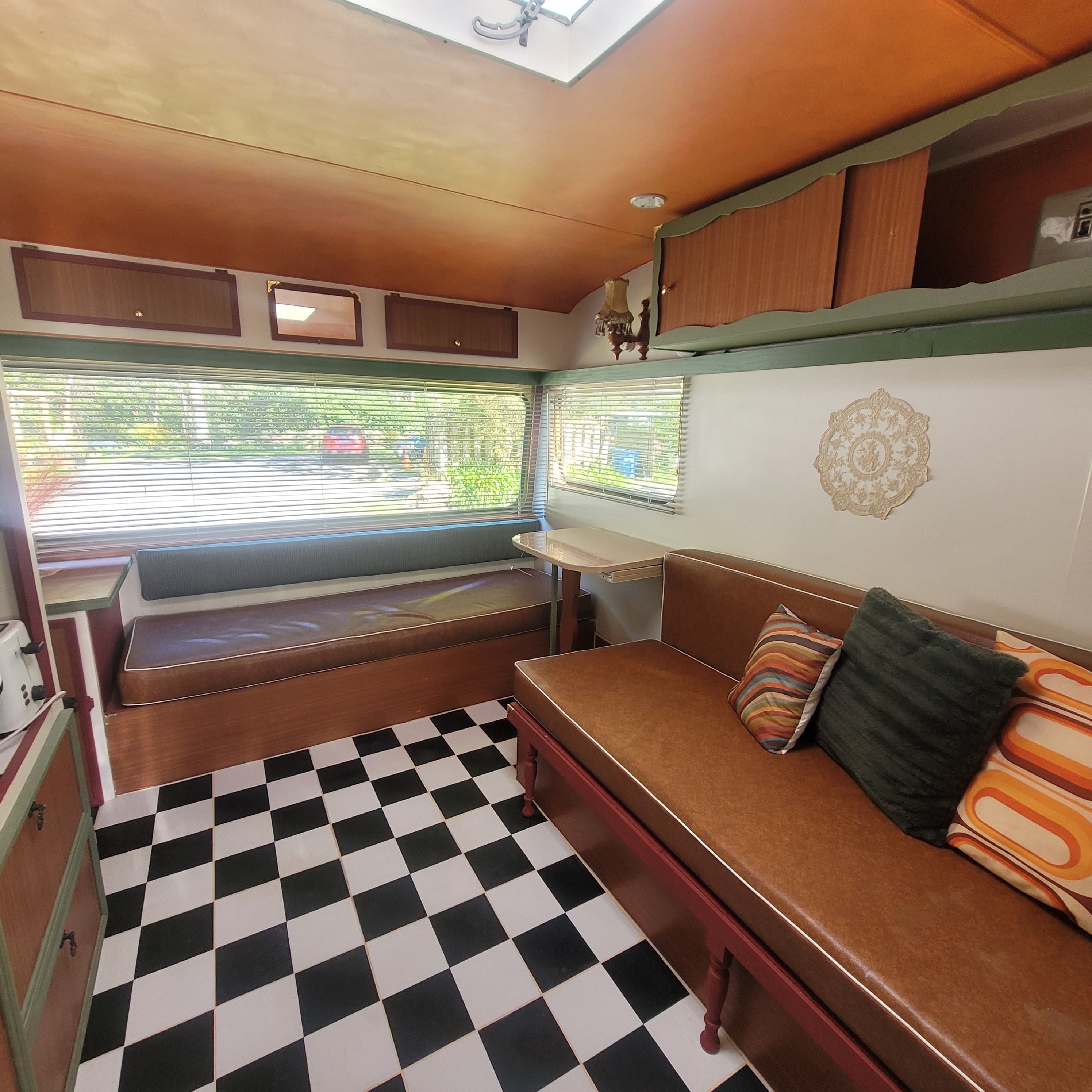 Un intérieur de camping-car compact avec un sol à carreaux, un coin salon avec des coussins, une petite table et une fenêtre avec des stores.
