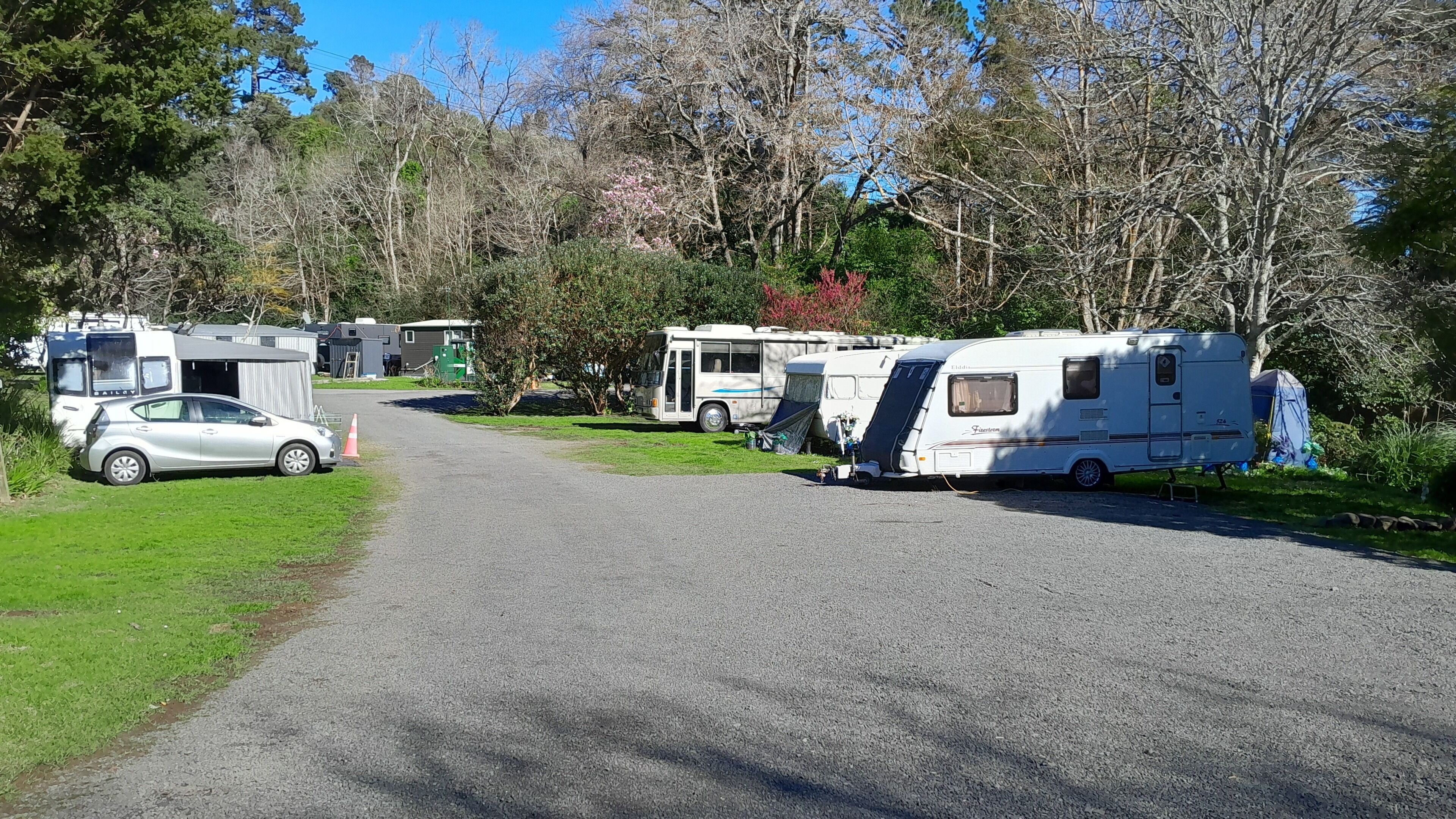 Un camping con vehículos recreativos y un coche aparcados en una carretera pavimentada, rodeado de árboles y follaje.