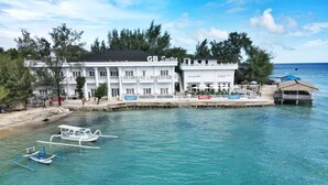 Un hotel frente a la playa con fachada blanca, llamado 'GB Suites', que cuenta con un muelle con barcos y una estructura de techo de paja.
