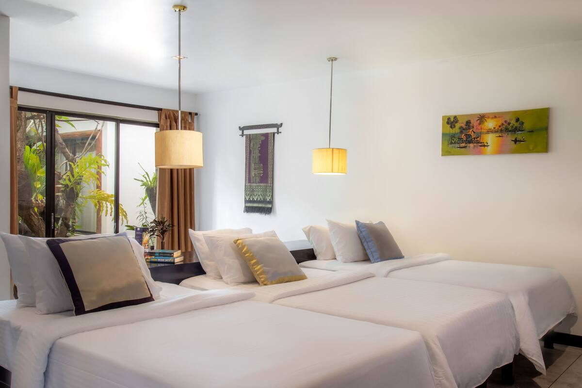A hotel room with two beds, a large window with a view of greenery, and a painting on the wall.