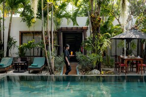 A pool area with lounge chairs, a table, and a covered seating area surrounded by tropical plants.