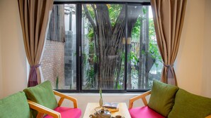 A room with a wooden table, two green cushioned chairs, a red sofa, a window with a view of a tree and greenery, and a brick wall. - Bunwin Siem Reap (Siem Reap)