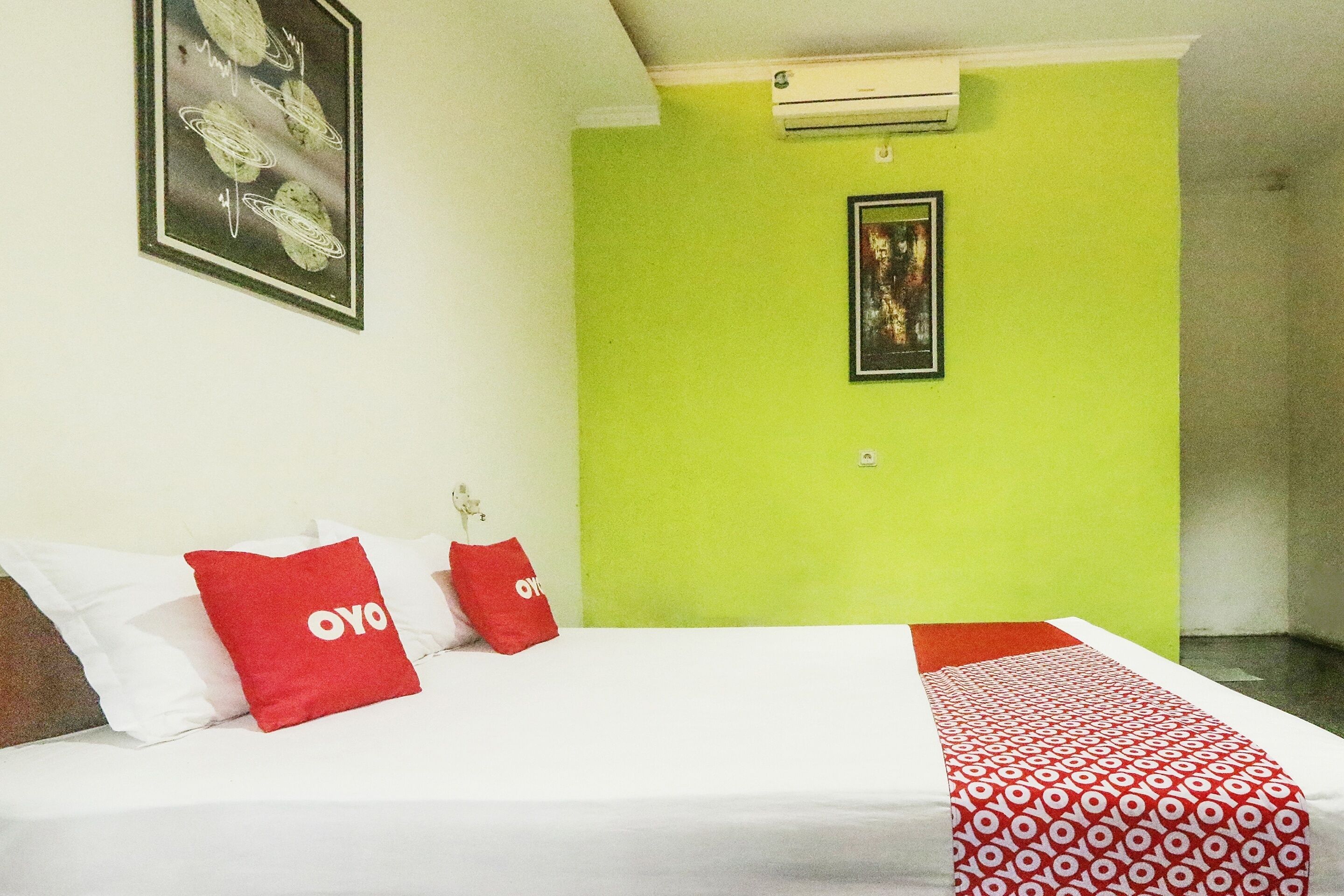 A hotel room with a bed, two red pillows with 'OYO' branding, a green wall, a framed picture, and an air conditioning unit.