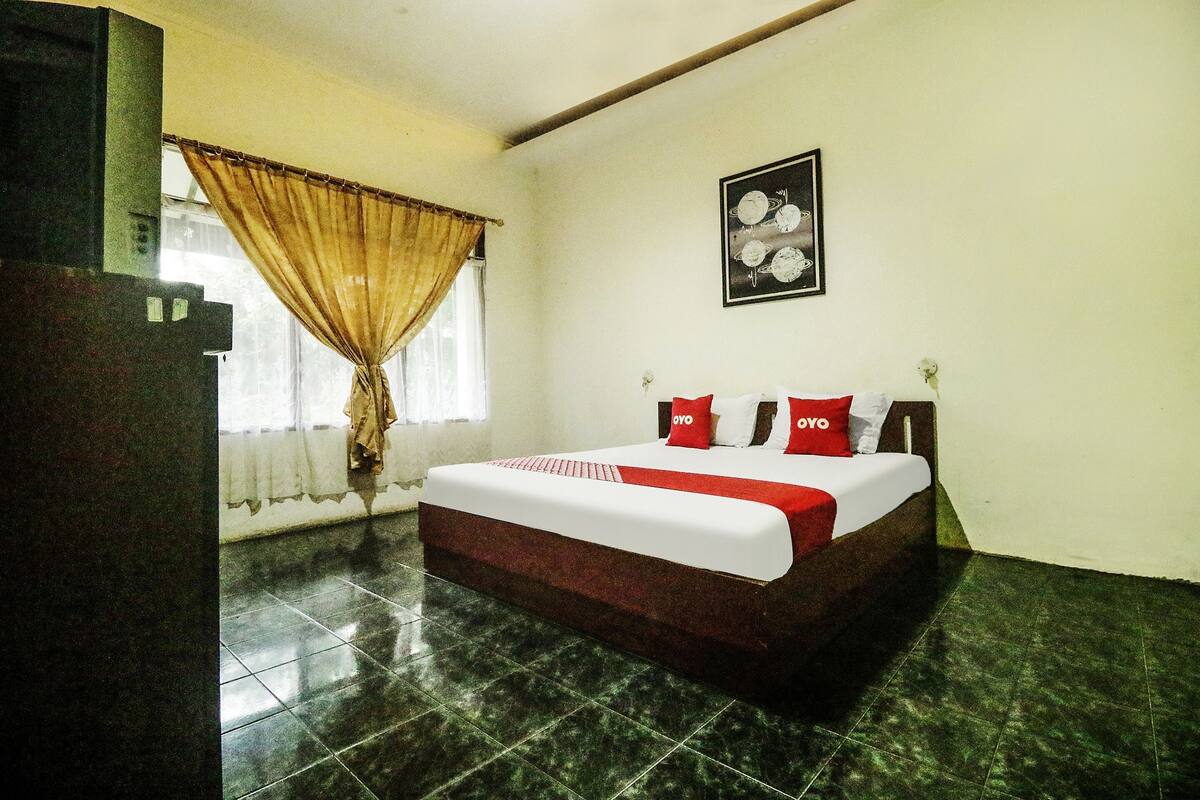 Une chambre d’hôtel avec un lit, une fenêtre avec des rideaux, un tableau encadré au mur et un sol carrelé.
