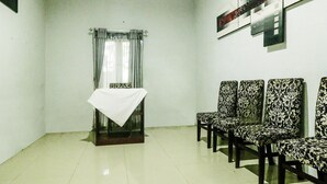 A waiting area with a table covered by a white cloth, a window with curtains, and a row of patterned chairs. - Hotel O Nyi Rindang Near Puskesmas Jekan Raya (Palangkaraya)